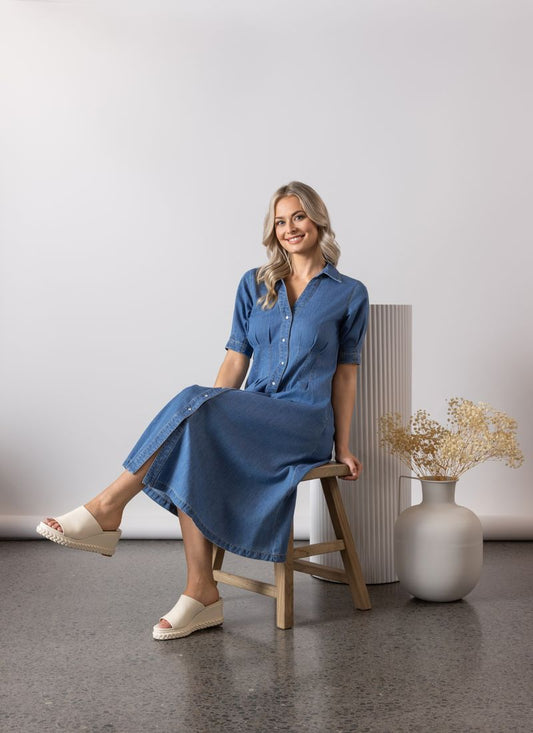 Woman wearing the Vassalli Shaped blue dress sitting in a minimalistic room with a white wall and wooden stool.