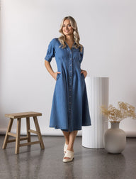 Woman wearing the Vassalli Shaped blue dress standing in a minimalistic room with a white wall and wooden stool.