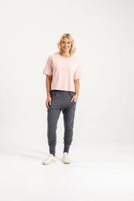 Lady wearing Home-Lee Apartment Pants in Graphite with Blossom X, pink Tee, white sneakers and a white background