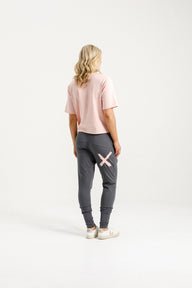 Lady wearing Home-Lee Apartment Pants in Graphite with Blossom X, pink Tee, white sneakers and a white background