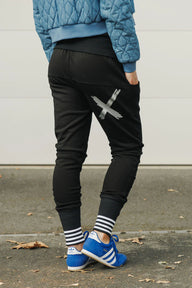 Person wearing black jogger pants with a large 'X' design, blue jacket, and blue sneakers on a concrete surface.