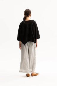 A woman shown from the back wearing a loose black top with eyelet embroidery, light gray wide-leg pants, and brown flat shoes, photographed against a plain white studio background.