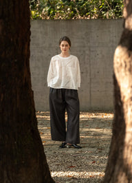 A woman stands outdoors between two large tree trunks, wearing a white textured knit long-sleeve top and loose black wide-leg pants, with a concrete wall and gravel ground behind her.