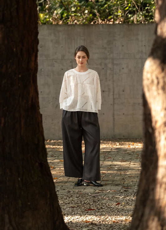 A woman stands outdoors between two large tree trunks, wearing a white textured knit long-sleeve top and loose black wide-leg pants, with a concrete wall and gravel ground behind her.