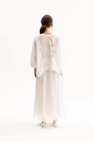A woman viewed from behind wearing a white crochet knit cardigan with geometric openwork patterns over a long white skirt, set against a plain white studio background.