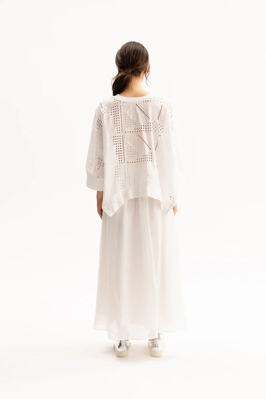 A woman viewed from behind wearing a white crochet knit cardigan with geometric openwork patterns over a long white skirt, set against a plain white studio background.