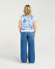 A woman shown from the back wearing a white short-sleeve top with a blue floral pattern and wide-leg blue jeans, standing against a light gray studio background.