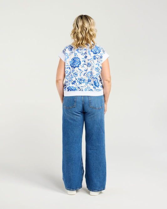 A woman shown from the back wearing a white short-sleeve top with a blue floral pattern and wide-leg blue jeans, standing against a light gray studio background.