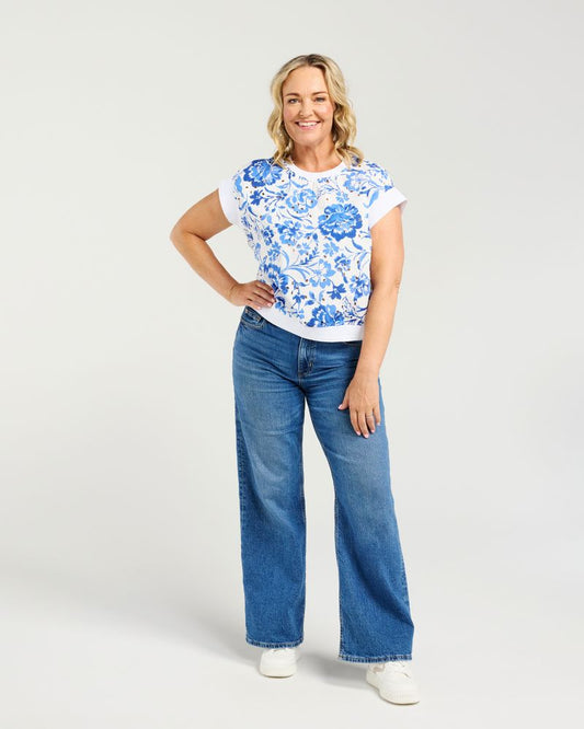 A woman modeling a white short-sleeve t-shirt with a blue floral pattern, paired with wide-leg blue jeans and white sneakers, against a light gray studio background.