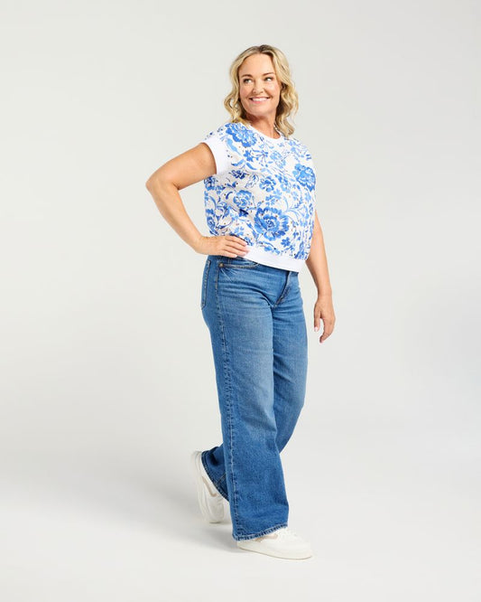 A woman modeling a short-sleeve white t-shirt with a blue floral pattern, paired with wide-leg blue jeans and white sneakers, against a light gray studio background.