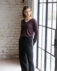 Woman wearing a purple sweater and dark pants standing in a room with a brick wall and large window.