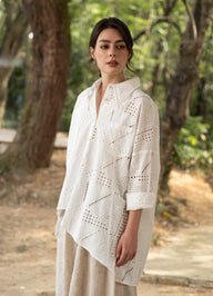 A woman wearing a white eyelet crochet shirt jacket with three-quarter sleeves and a high collar, paired with beige linen trousers, standing in a sunlit outdoor forest setting.