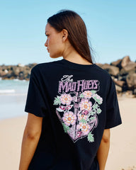 A woman standing on a beach, shown from the back, wearing a black t-shirt with a large graphic print featuring the text 'The Mad Hueys' and a floral heart design.
