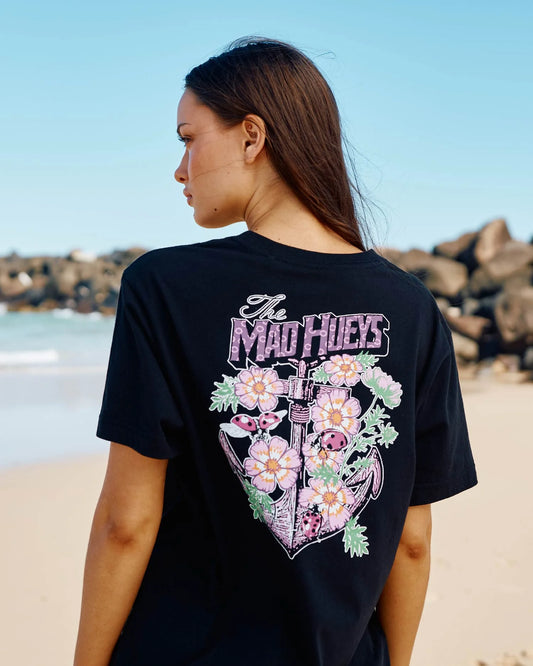 A woman standing on a beach, shown from the back, wearing a black t-shirt with a large graphic print featuring the text 'The Mad Hueys' and a floral heart design.