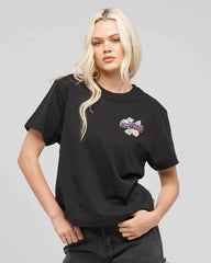 A blonde model wearing a black short-sleeve t-shirt with a colorful graphic on the left chest that reads mad hueys' in purple with pink flowers and green leaves, paired with dark gray jeans against a solid gray studio background.