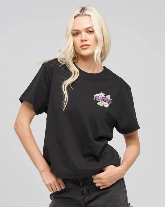 A blonde model wearing a black short-sleeve t-shirt with a colorful graphic on the left chest that reads mad hueys' in purple with pink flowers and green leaves, paired with dark gray jeans against a solid gray studio background.