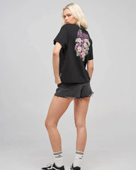 A blonde model wearing a black graphic t-shirt with a large floral print on the back, paired with dark gray denim shorts, white socks, and black sneakers, photographed against a light gray studio background.
