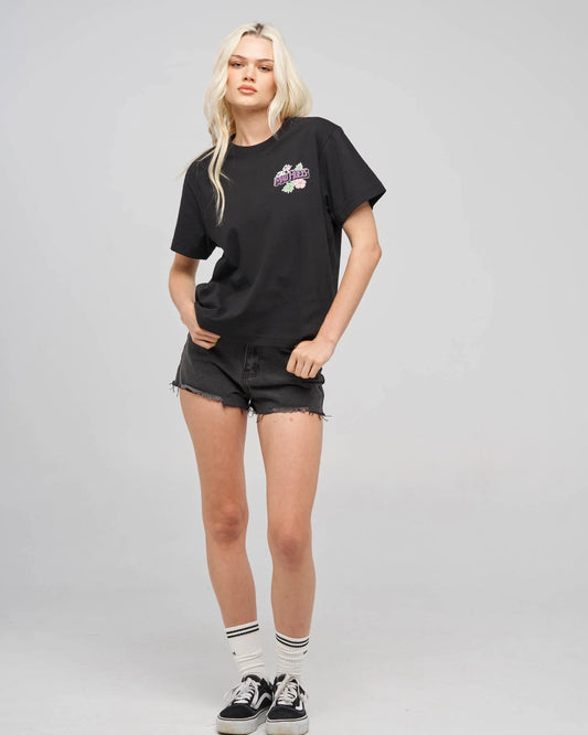 A blonde model wearing a black short-sleeve graphic t-shirt with a small colorful chest print, black denim shorts, white socks, and black sneakers, standing against a light gray studio background.