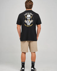 A man viewed from behind wearing a black graphic t-shirt with a large yellow and white skull design on the back, paired with beige shorts, black socks, and white sneakers, against a light gray studio background.