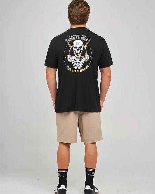 A man viewed from behind wearing a black graphic t-shirt with a large yellow and white skull design on the back, paired with beige shorts, black socks, and white sneakers, against a light gray studio background.