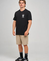 A man modeling a black short-sleeve t-shirt with a small white chest graphic, paired with beige shorts, black socks, and black sneakers, against a light gray studio background.