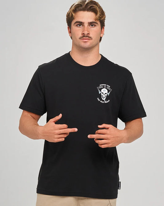 A man wearing a black short-sleeve t-shirt with a white skull graphic on the left chest, paired with beige pants, against a solid gray studio background.