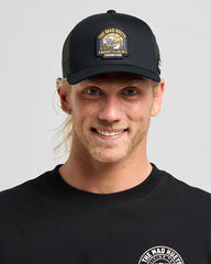 Man wearing a black cap with a logo and matching black shirt on a gray background