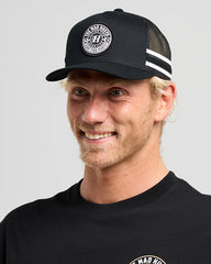 Man wearing a black cap with a logo and a matching black shirt on a gray background