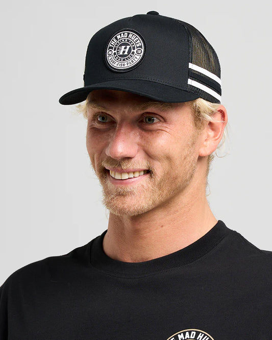 Man wearing a black cap with a logo and a matching black shirt on a gray background