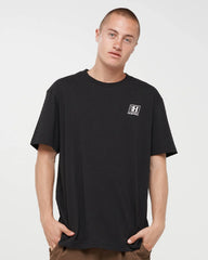 A man wearing a black short-sleeve crewneck t-shirt with a small white graphic logo on the left chest, posed against a light gray studio background.