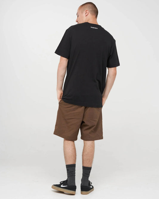 A man wearing a black t-shirt, brown shorts, black socks, and black sneakers, photographed from behind against a light gray studio background.
