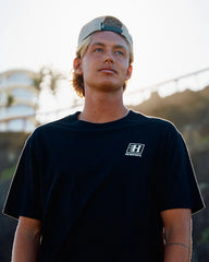 A man wearing a black t-shirt with a white 'The Brand of It' logo on the left chest, paired with a beige cap, photographed outdoors against a bright, sunlit background.