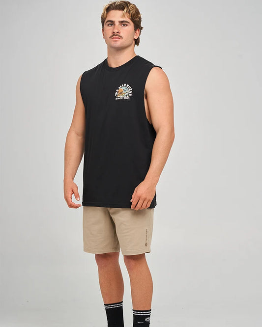 A man modeling a black sleeveless muscle tank top with a small multicolor graphic on the left chest, paired with beige shorts and black socks, against a light gray studio background.