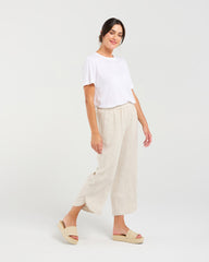 A woman modeling a white short-sleeve t-shirt and light beige linen wide-leg cropped pants with beige platform slide sandals, set against a plain light gray studio background.