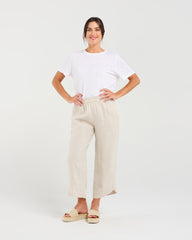 A woman modeling a white short-sleeve crewneck t-shirt and light beige linen wide-leg cropped pants with beige slide sandals, standing against a light gray studio background.