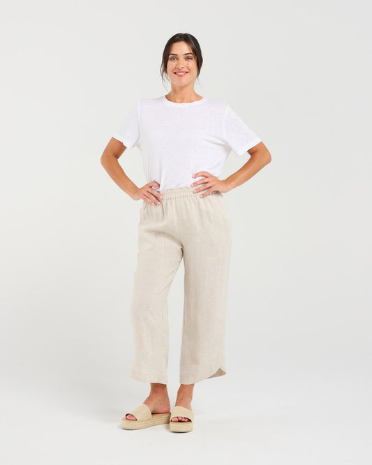 A woman modeling a white short-sleeve crewneck t-shirt and light beige linen wide-leg cropped pants with beige slide sandals, standing against a light gray studio background.