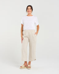 A woman modeling a white short-sleeve crewneck t-shirt and light beige linen wide-leg cropped pants with side pockets, paired with beige platform slide sandals, against a solid light gray studio background.