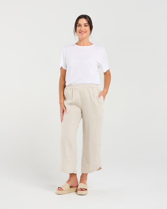 A woman modeling a white short-sleeve crewneck t-shirt and light beige linen wide-leg cropped pants with side pockets, paired with beige platform slide sandals, against a solid light gray studio background.