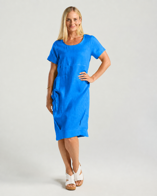 A woman modeling a bright blue short-sleeve linen dress with side pockets, paired with white wedge sandals, against a light gray studio background.