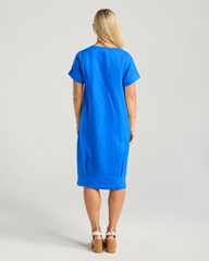 A woman viewed from behind wearing a bright blue short-sleeve linen dress, standing against a light gray studio background.