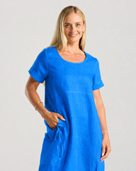A woman modeling a bright blue short-sleeve linen dress with a round neckline and a side pocket, photographed against a light gray studio background.