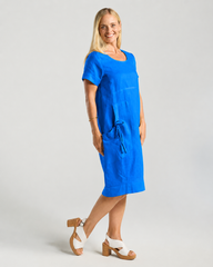 A woman modeling a bright blue linen dress with short sleeves, a round neckline, and a drawstring detail at the waist, paired with white and brown block-heel sandals against a light gray studio background.