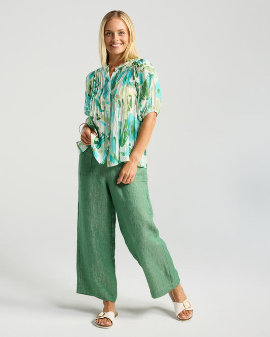Woman wearing a green and white patterned blouse with green pants on a light gray background