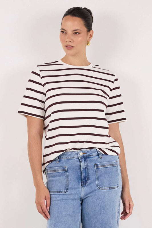 A woman wearing a short-sleeve crewneck t-shirt with dark brown horizontal stripes on an off-white background, paired with light blue jeans, against a plain light gray studio background.