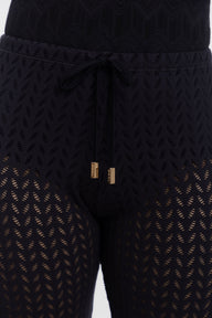 Close-up of black textured knit leggings with a drawstring waist and gold-toned hardware, shown on a model against a light gray background.
