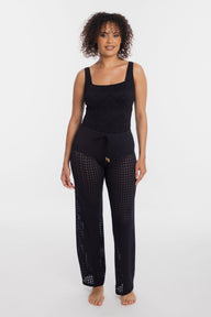 A woman modeling a black knit lounge set consisting of a sleeveless top and matching drawstring pants with open-knit leg panels, photographed against a plain light gray studio background.