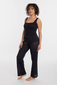 A woman modeling a black sleeveless jumpsuit with a textured, open-knit pattern on the pants, standing against a plain white studio background.