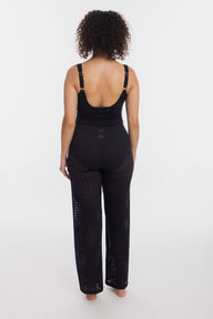 A woman viewed from the back wearing a black open-knit jumpsuit with a low open back and wide legs, photographed against a plain light gray studio background.