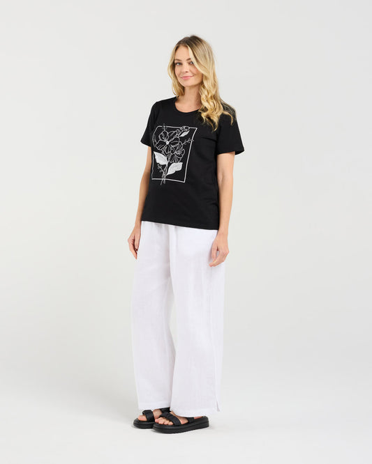 Woman wearing a black t-shirt with a graphic design and white pants on a light gray background