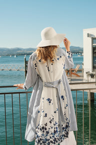 Woman in a floral dress and wide-brimmed hat standing by a waterfront with mountains in the background.
